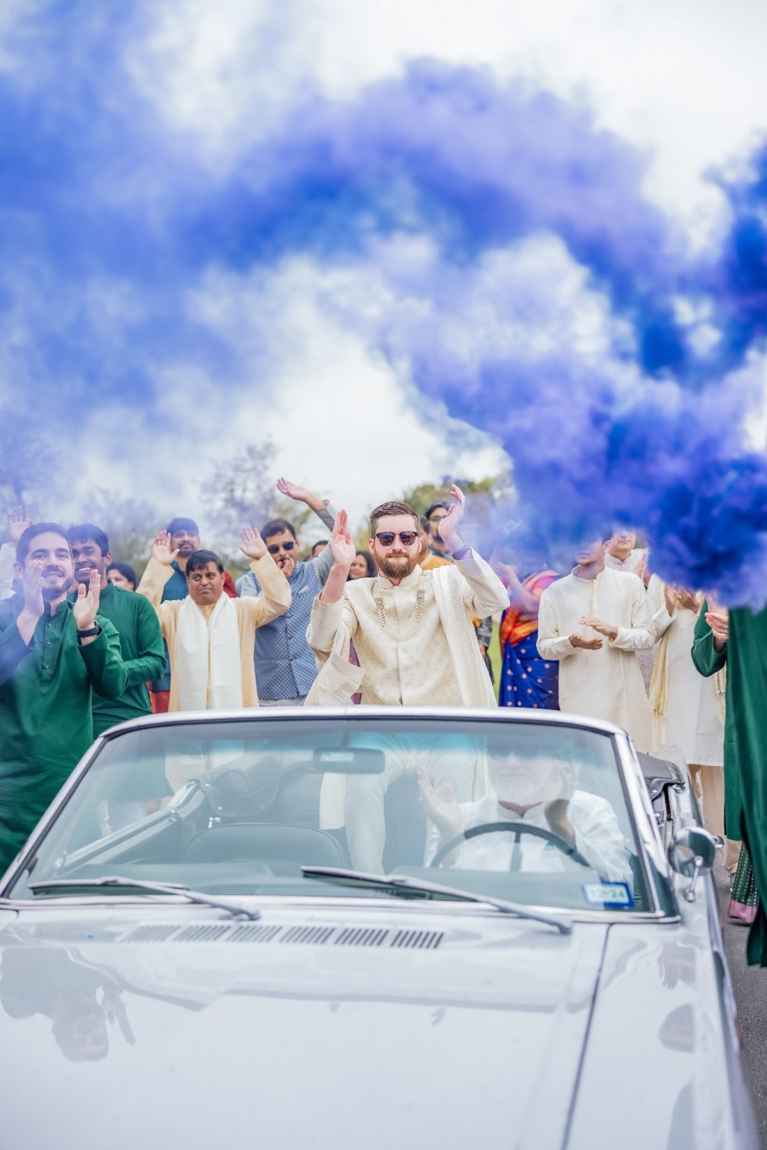 Groom in a convertible surrounded by blue smoke and clapping friends during the baraat