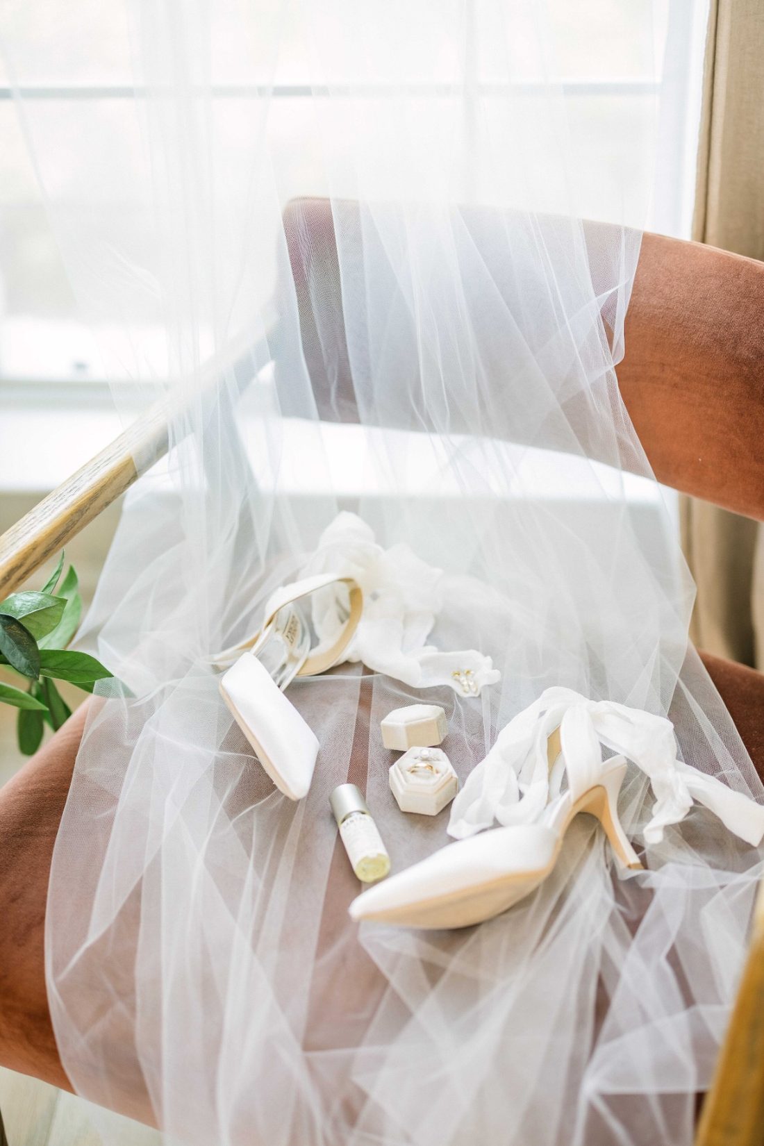 Bridal heels, ring box, and perfume nestled in a flowing veil on a velvet chair