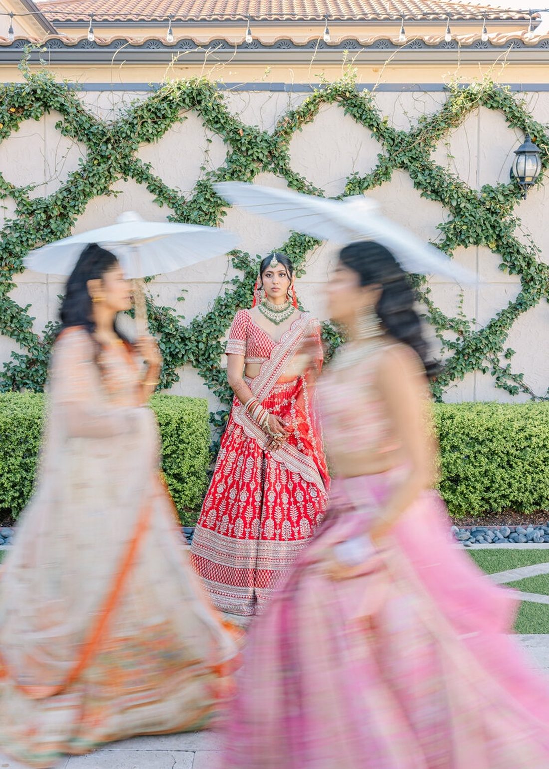 Bride in red lehenga framed by friends in flowing motion blur near a vine wall