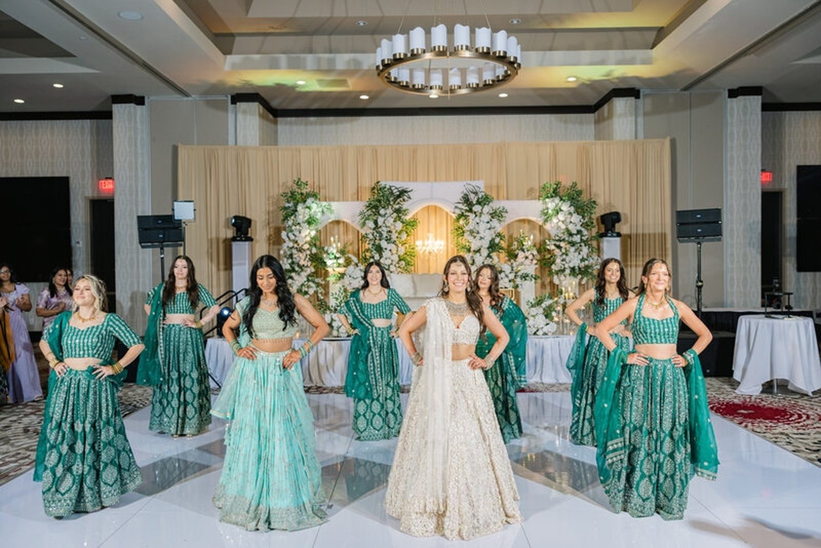 Bride and bridesmaids posed in coordinated emerald and teal lehengas on a ballroom dance floor